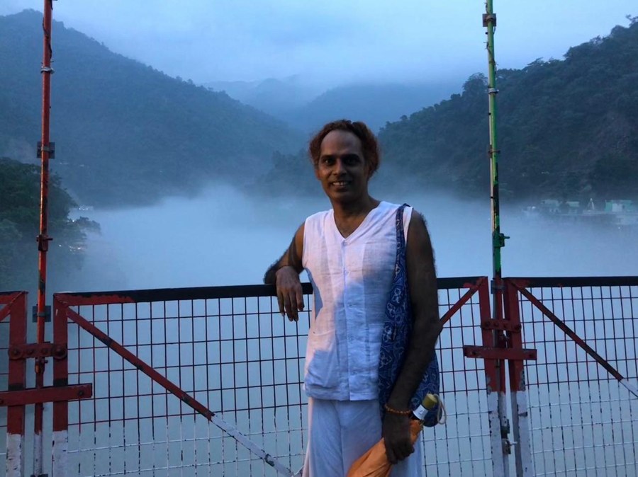 Ramabuddha standing on a railing above the misty river in Rishikesh, with green hills behind him