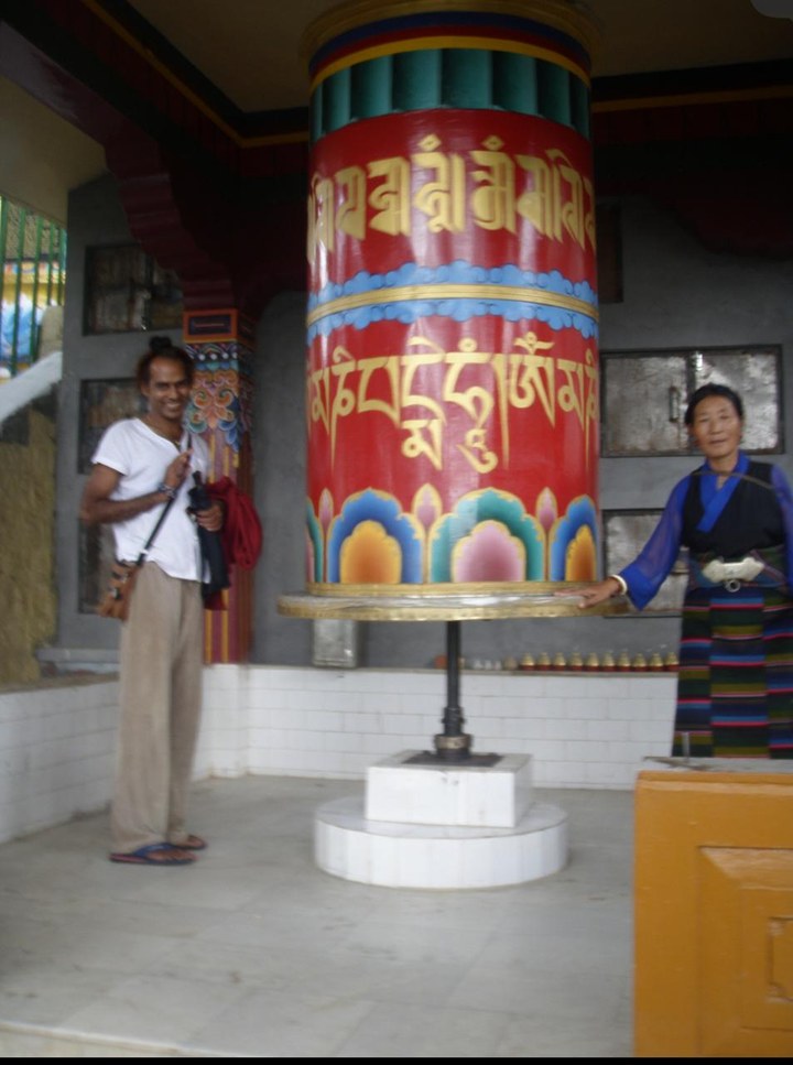 Ramabuddha as a young man, smiling beside a large painted prayer wheel